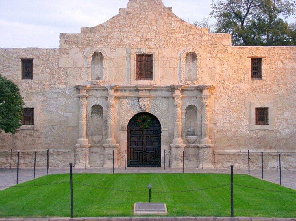 The limestone facade of the Alamo Church in San Antonio with its arched doorway, carved columns and green lawn