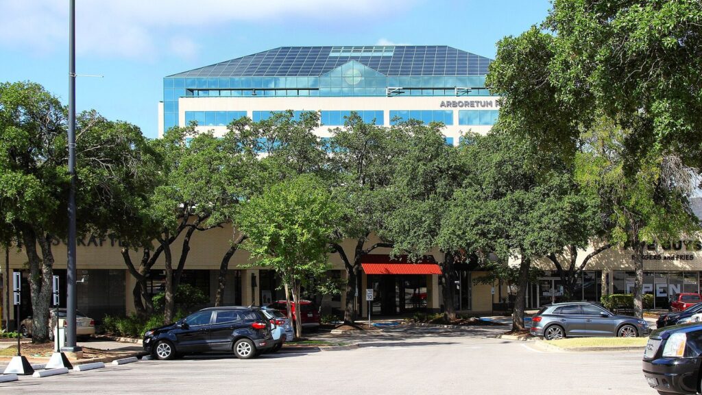 Low shop buildings and a parking lot shaded by live oaks at The Arboretum in Northwest Austin