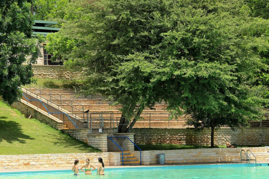 Swimmers standing in the clear water of Deep Eddy Pool below its limestone stairs, retaining walls and shade trees