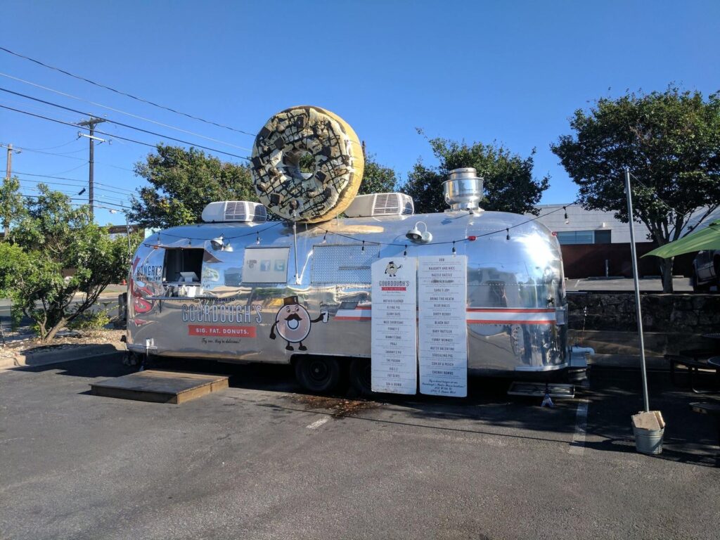 A silver Airstream food trailer with a giant doughnut sculpture on its roof, parked in a sunny Austin lot
