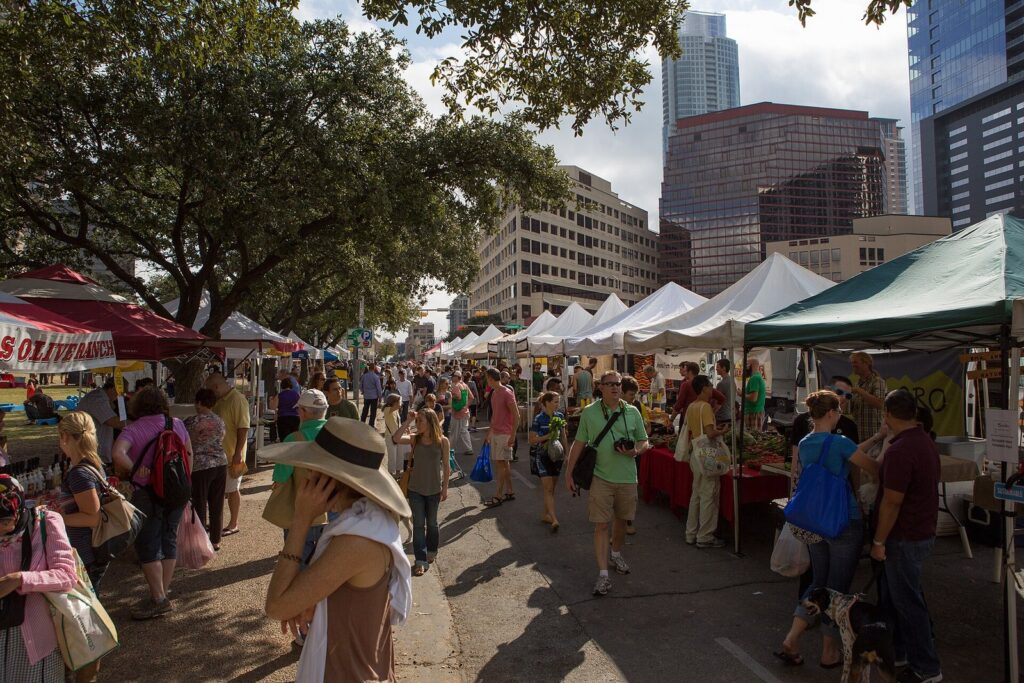 Shoppers walking between white market tents under trees, with downtown Austin office towers behind