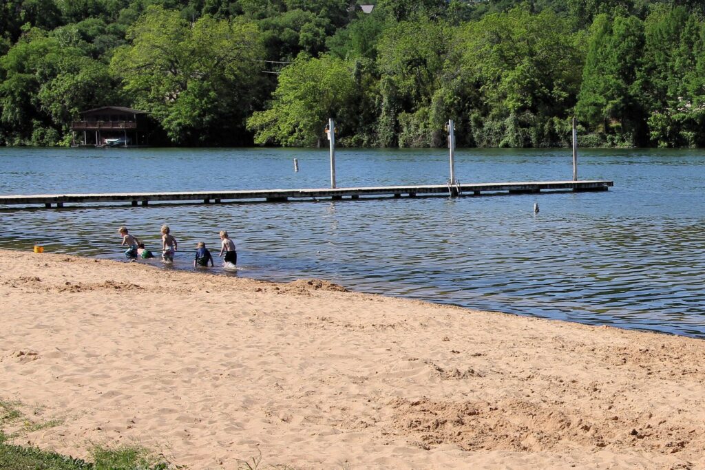 Kids wading off the sandy beach at Emma Long Metropolitan Park, one of the Austin swimming holes and lakes closest to the city, with a pier on Lake Austin behind