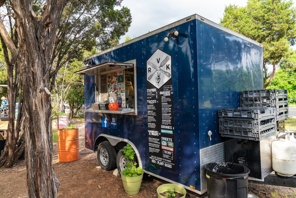 A navy-blue food trailer with a menu board and service window under cedar trees at a South Austin food park