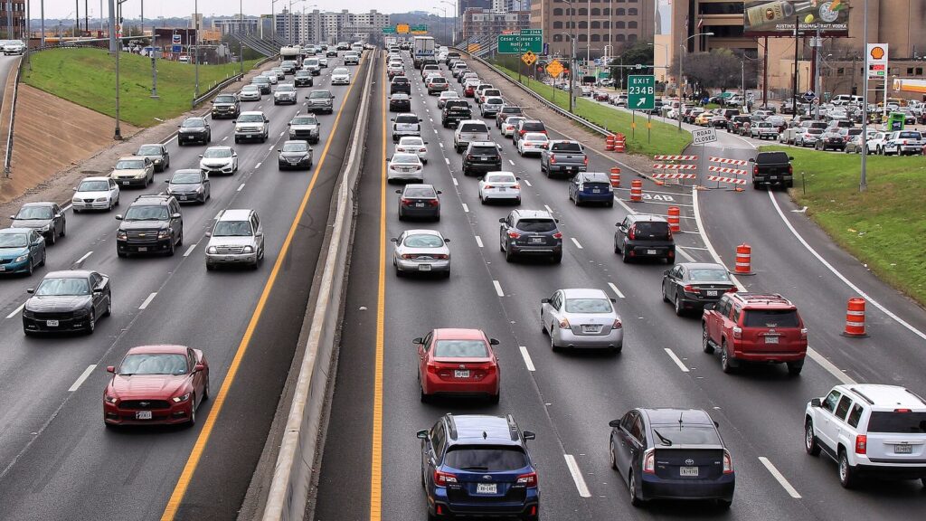 Several lanes of cars on Interstate 35 through downtown Austin on a weekend, with orange construction barrels and an exit sign