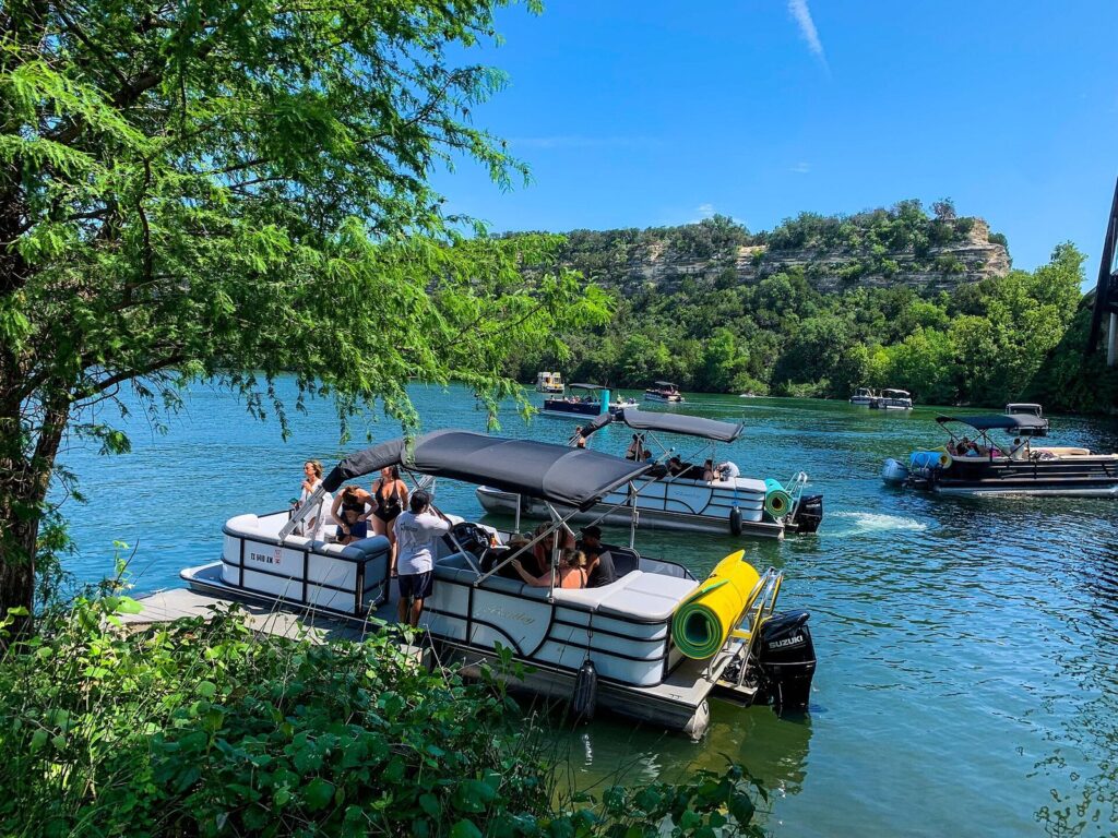 Pontoon boats loading at a dock on Lake Austin, with green bluffs and the Pennybacker Bridge in the distance