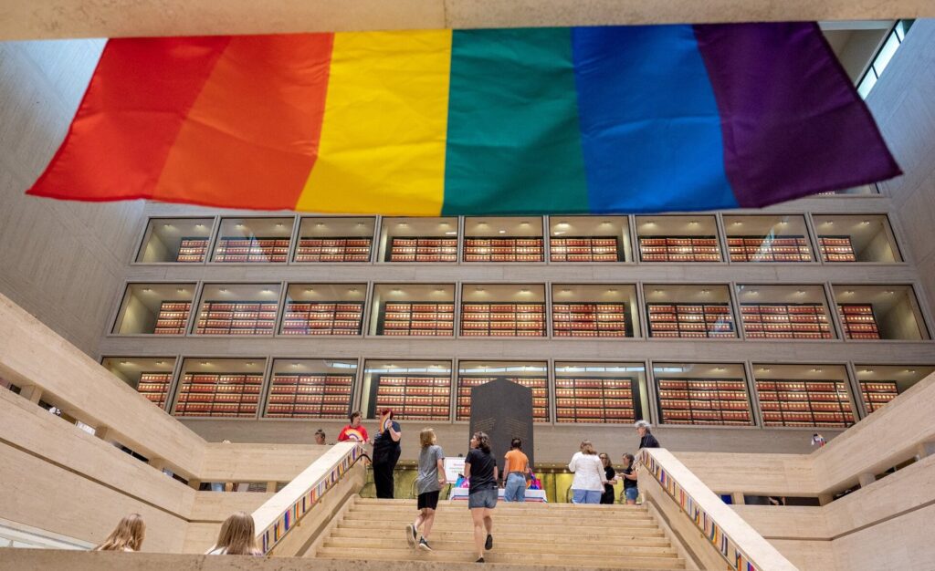 A large rainbow Pride flag hanging in the LBJ Presidential Library's Great Hall above visitors on the stairs and the archive stacks