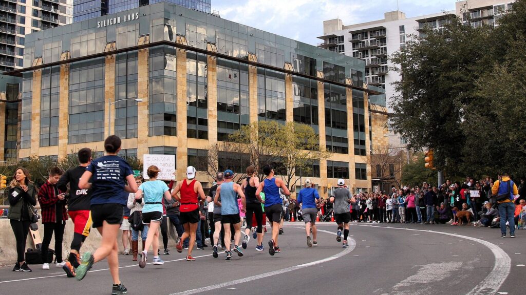 Runners and spectators on a downtown Austin street during the Austin Marathon, with glass office towers behind