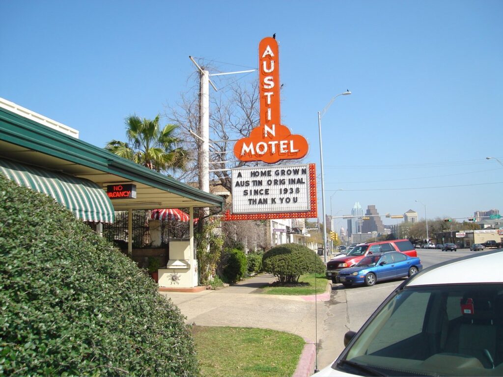 The tall orange Austin Motel neon sign on South Congress Avenue, reading a home grown Austin original since 1938, with downtown towers in the distance, a landmark among the best hotels on South Congress Austin