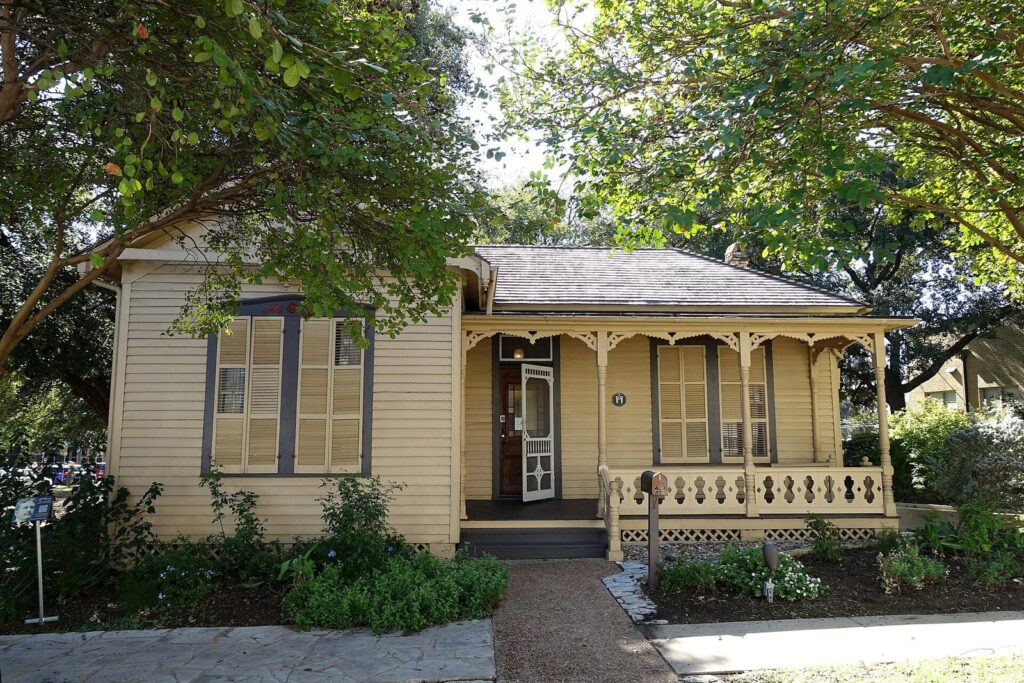 The small cream-colored Queen Anne cottage of the O. Henry Museum in Brush Square, with a front porch and shade trees