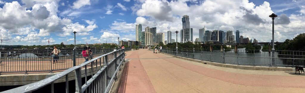 Walkers, cyclists, scooter riders and pedicabs getting around Austin on the Pfluger Pedestrian Bridge over Lady Bird Lake, with the downtown skyline ahead