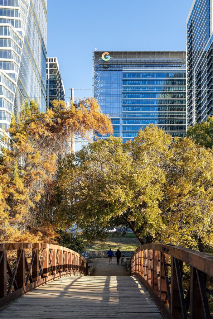 A wooden footbridge on the Shoal Creek Trail framed by autumn trees, with downtown Austin towers rising behind