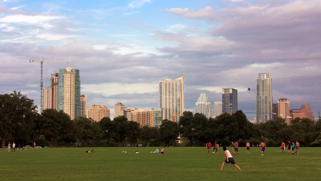 The downtown Austin skyline rising beyond the trees and open lawn of Zilker Park, with people playing on the grass