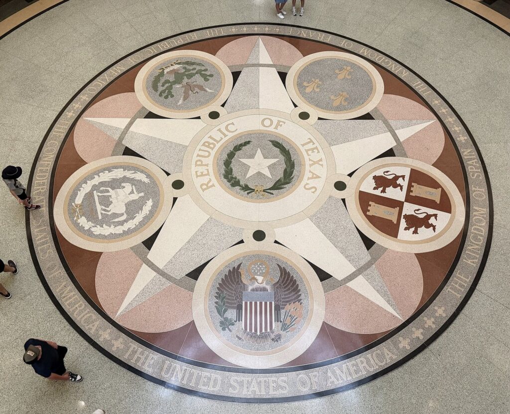 The star-patterned terrazzo floor of the Texas State Capitol rotunda seen from the balconies above, with visitors crossing it