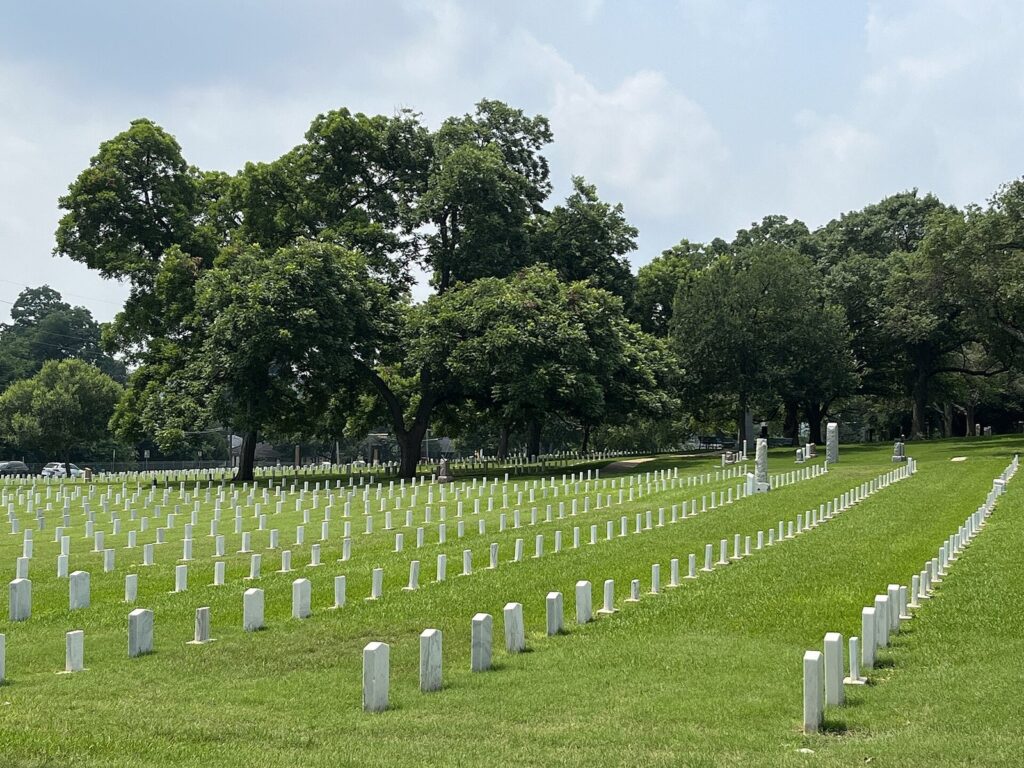 Rows of white headstones curving across green lawns under large shade trees at the Texas State Cemetery in East Austin