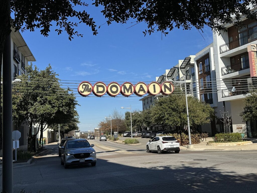A retro-style sign spelling The Domain strung across a street between apartment buildings and trees in North Austin