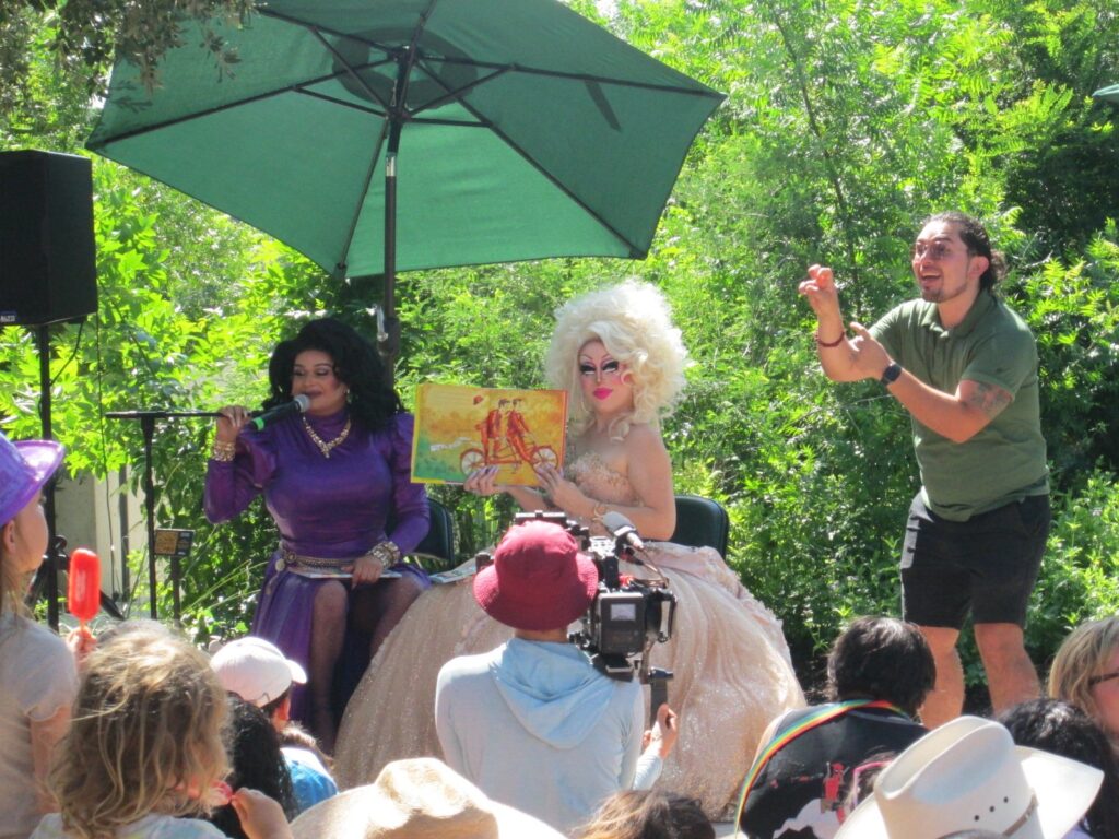 Two drag performers reading a picture book to families under trees at Waterloo Park during a Pride event, with an ASL interpreter nearby