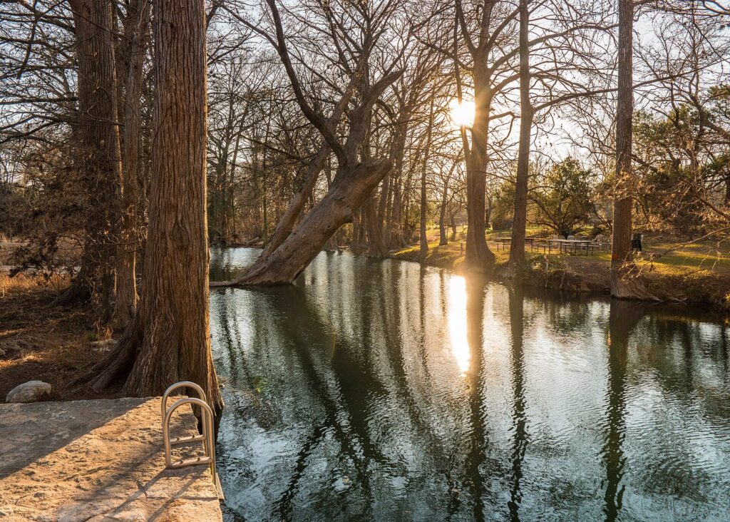 Sunlight through tall cypress trees reflected in the clear, still water of Blue Hole Regional Park in Wimberley