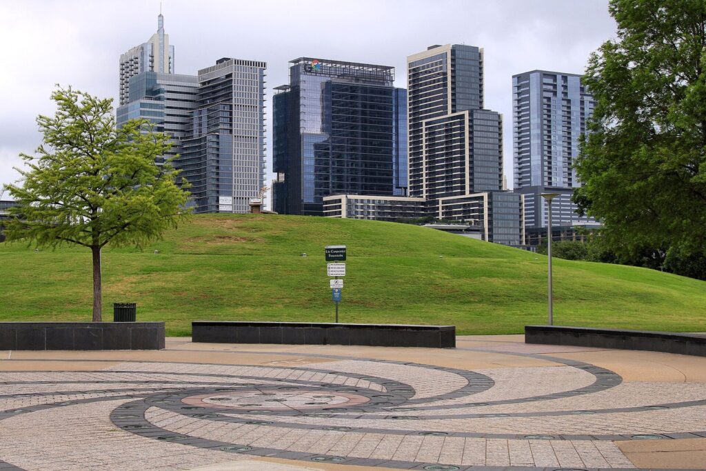 An open lawn and paved plaza at Butler Metro Park with downtown Austin towers rising behind the trees