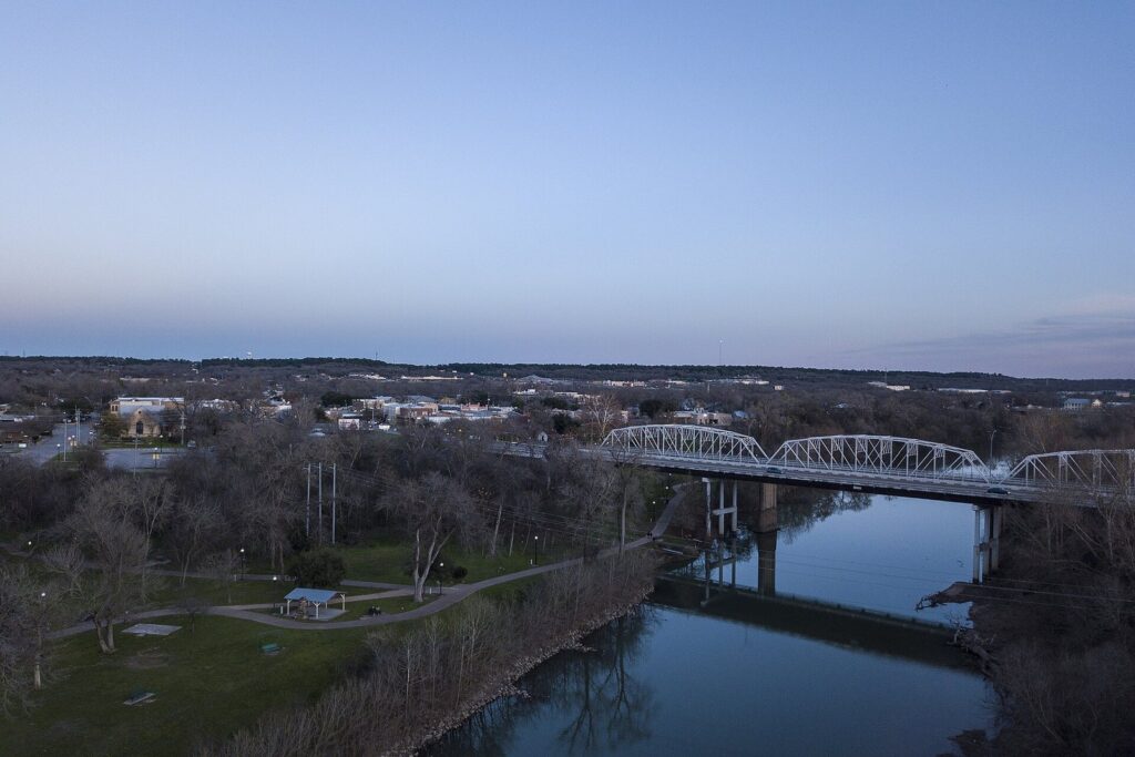 The Colorado River curving past a steel truss bridge and green parkland in Bastrop, Texas, at dusk