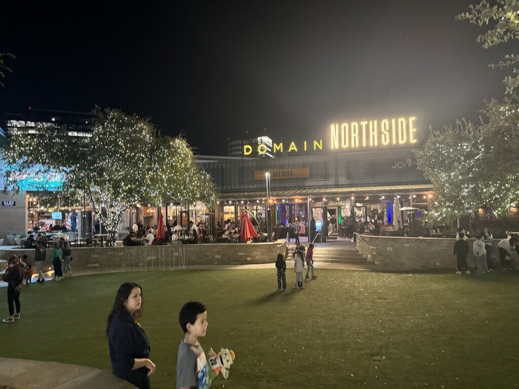 Domain Northside in North Austin at night, with a lit sign, restaurant patios and people on a lawn