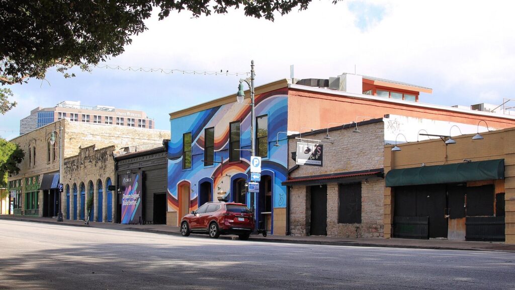 Low historic storefronts and a colorful mural along the 600 block of East Sixth Street in downtown Austin in daylight