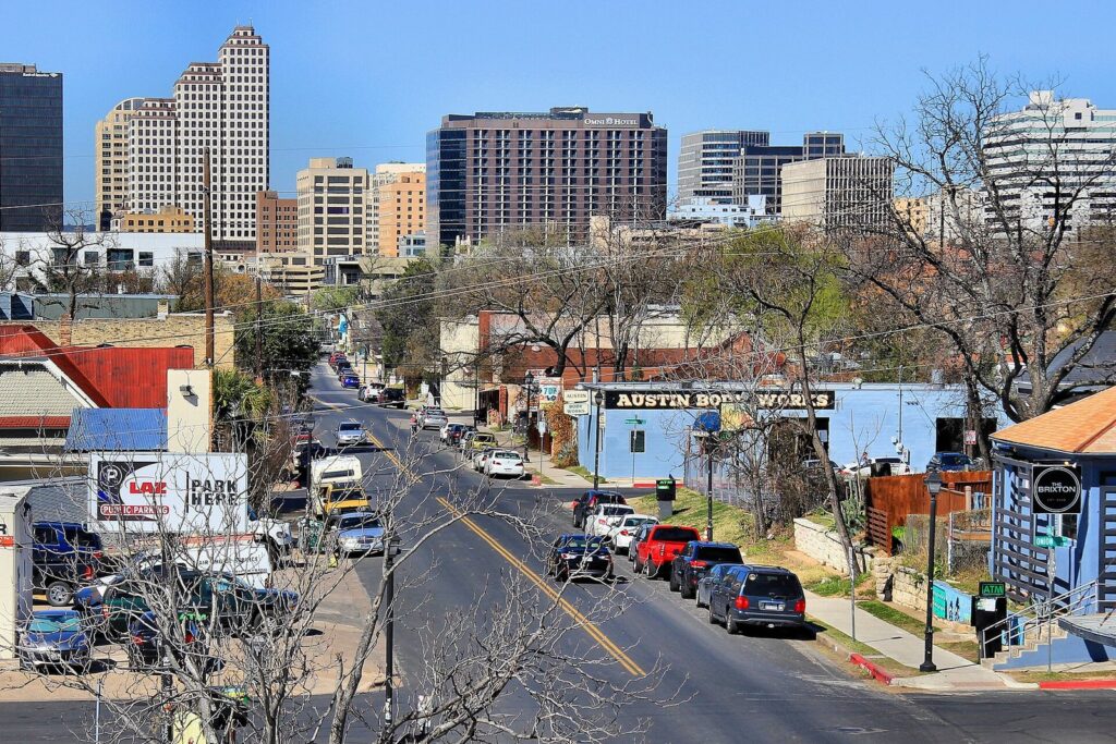 East Sixth Street in East Austin sloping toward the downtown skyline, with low shops and parked cars along the curb