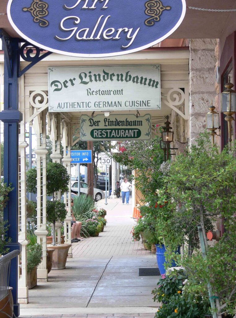 A shaded sidewalk on Fredericksburg's Main Street lined with shop signs, plants and old limestone buildings