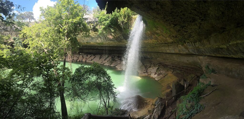 A thin waterfall pouring from a limestone overhang into the green water of Hamilton Pool, surrounded by trees