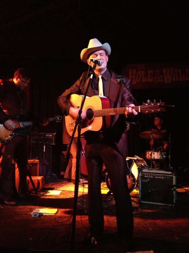 A singer in a cowboy hat playing acoustic guitar with his band on the small stage of the Hole in the Wall