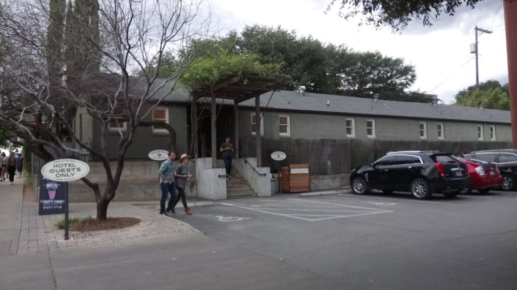 The low, gray bungalow buildings and gravel-edged entrance of Hotel San José on South Congress Avenue, with a hotel guests only sign by the parking lot