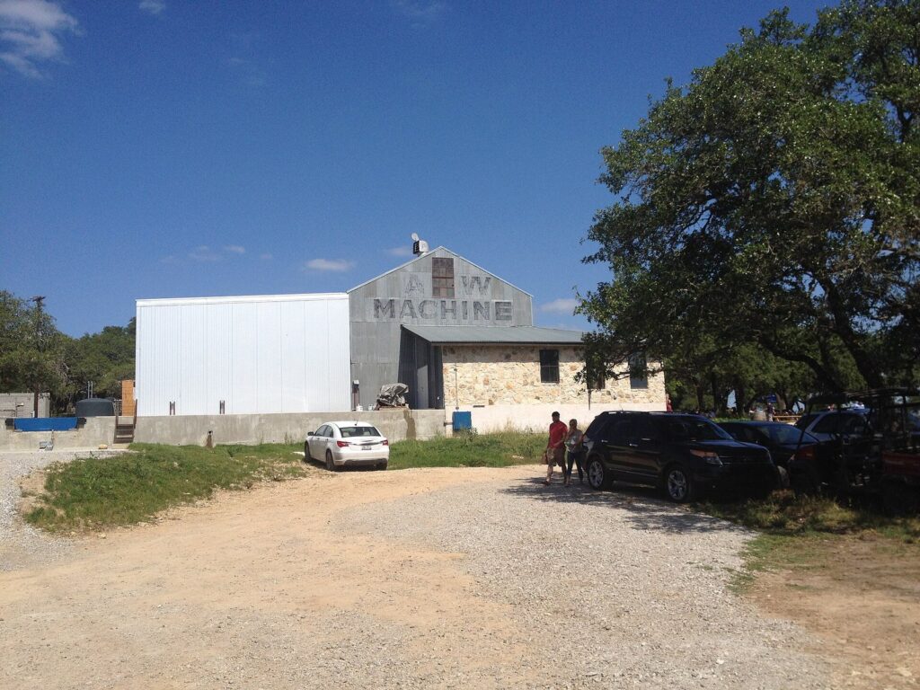 Jester King Brewery's metal-sided brewhouse and stone building beside a gravel parking area under a large oak in the Texas Hill Country