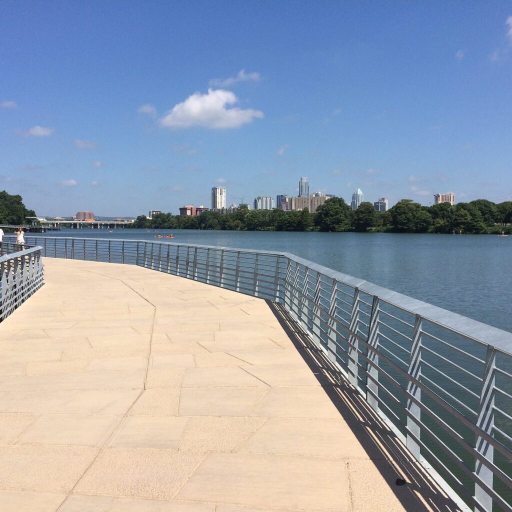 The wide pale boardwalk of the Butler Trail curving over Lady Bird Lake with metal railings and the downtown skyline ahead, part of this accessible Austin travel guide