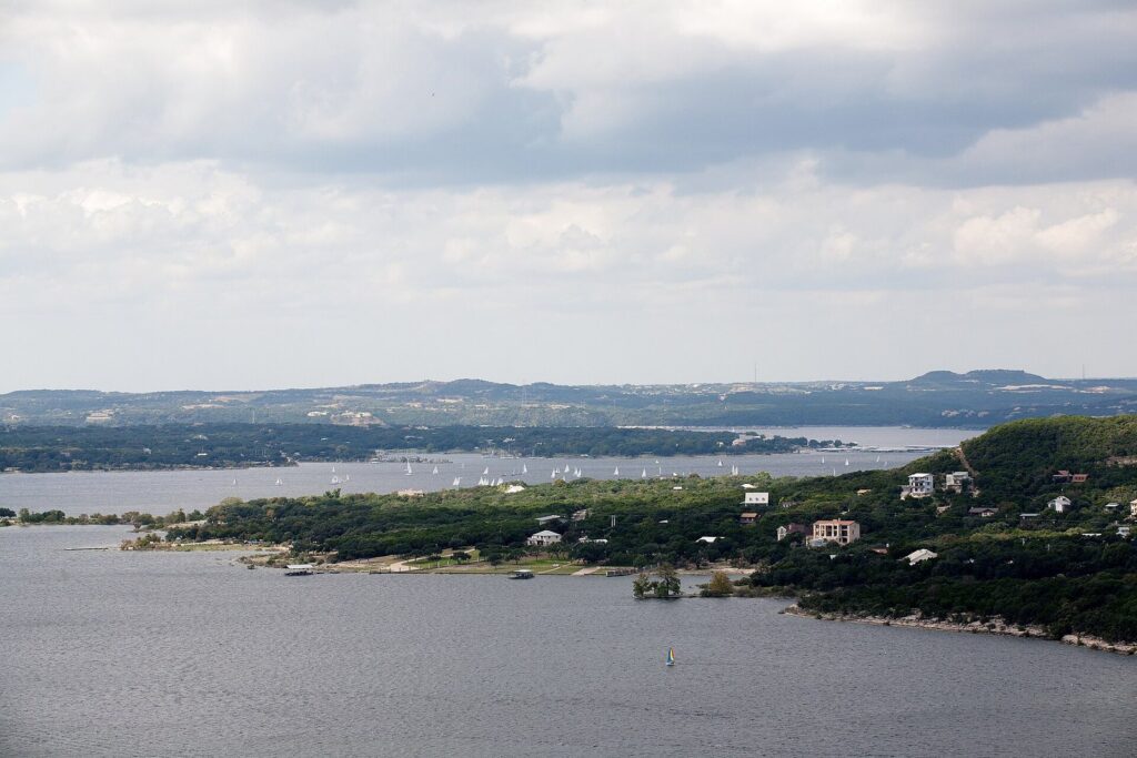 Lake Travis filled to the shoreline, with sailboats in the distance and wooded hills and houses along the far bank