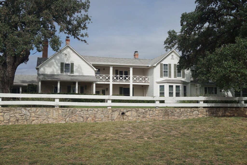 The white two-story Texas White House at the LBJ Ranch, with a stone wall, a white fence and large live oaks