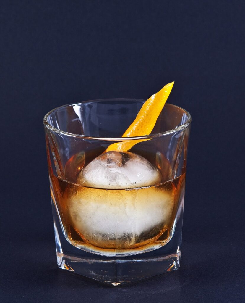 An Old Fashioned cocktail with a large clear ice sphere and a twist of orange peel against a dark blue background