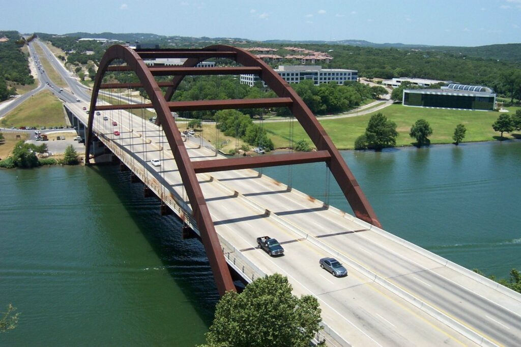 The rust-colored arch of the Pennybacker Bridge carrying Loop 360 traffic over Lake Austin, with wooded hills beyond