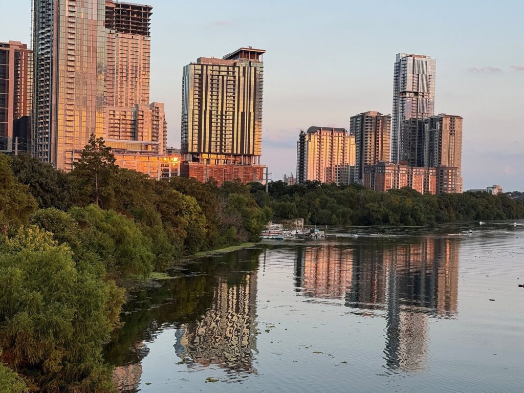 Rainey Street's high-rise towers glowing at sunset beside Lady Bird Lake, with trees along the shore and reflections in the water
