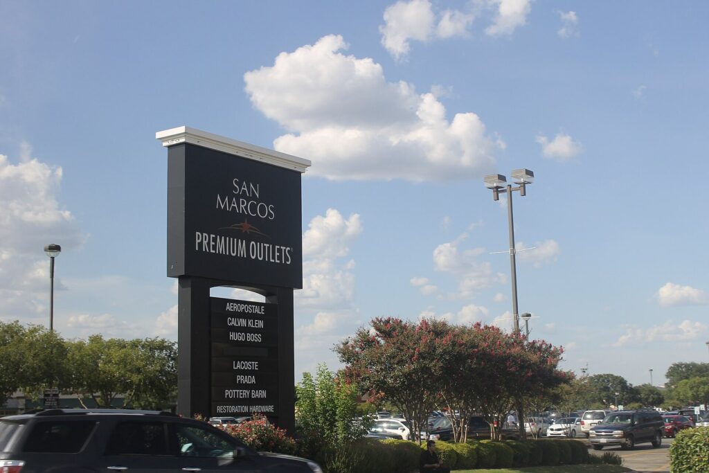 The tall San Marcos Premium Outlets sign listing brand names beside a parking lot under a summer sky