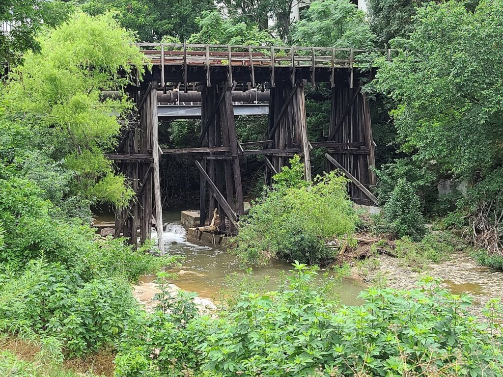 A weathered timber railroad trestle over Shoal Creek, framed by green trees and brush