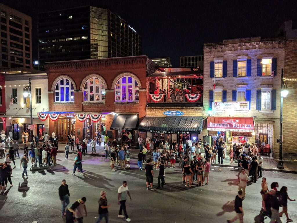 Crowds walking in the street among lit bars and historic buildings on Sixth Street in Austin at night, where some of the best cocktail bars in Austin hide behind quiet doors