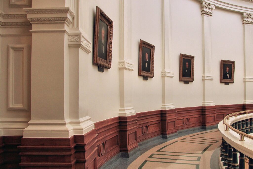 Framed portraits of Texas governors on the curved walls of the Texas State Capitol rotunda gallery, with a wooden railing in the foreground