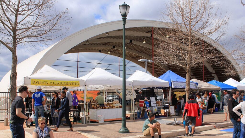 Vendor tents and shoppers under a curved white shade structure at the Texas Farmers' Market at Mueller in 2016