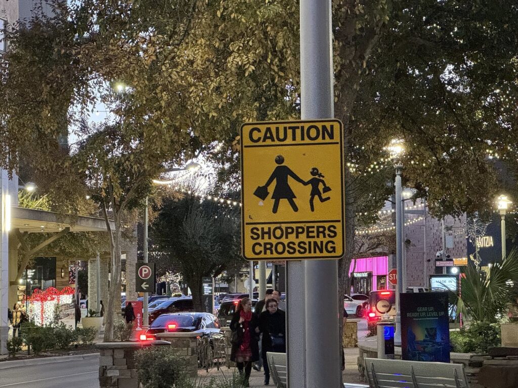 A yellow Caution Shoppers Crossing sign on a lamp post at The Domain in North Austin, with holiday lights and storefronts behind