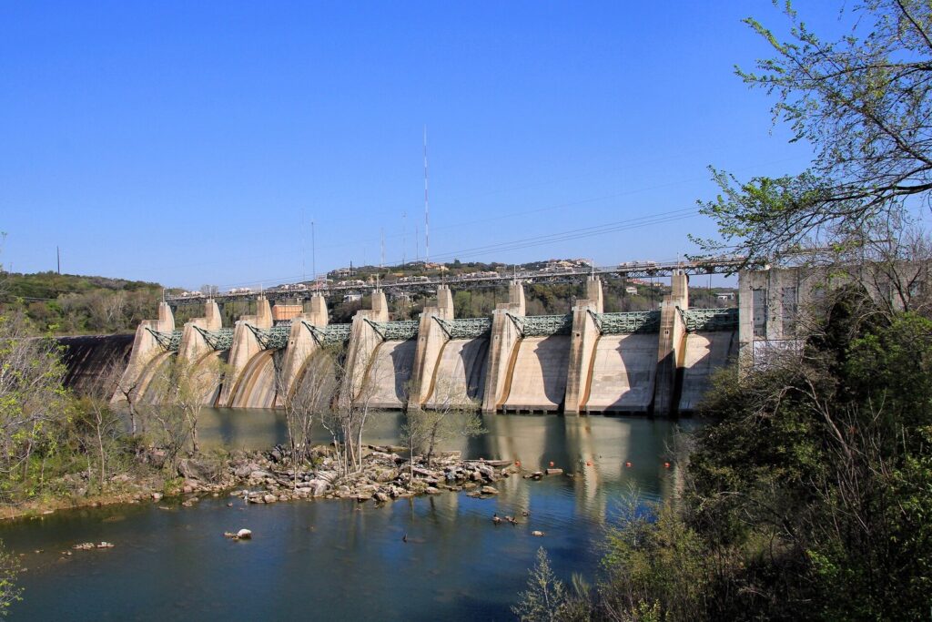 The concrete spillway gates of Tom Miller Dam above the Colorado River in West Austin, with rocks and trees in the foreground