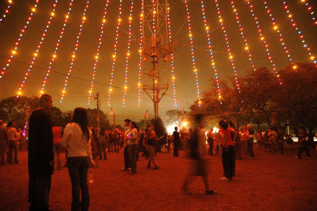 Strings of colored lights spiraling down from a tall moonlight tower over a crowd in Zilker Park at night