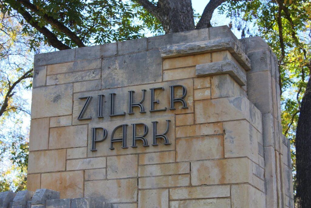 Art deco lettering reading Zilker Park on a limestone entrance sign under shade trees